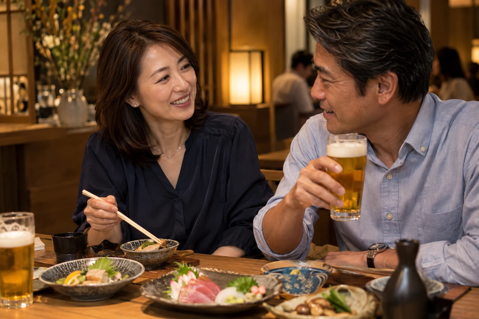 和食のお店で、紺色のブラウスの女性と男性が料理とビールを楽しむ。二人とも40代半ば。